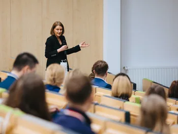 Kobieta w ciemnej marynarce przemawia do publiczności w sali wykładowej.