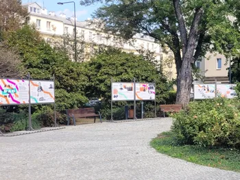 Tablice informacyjne w parku, otoczone zielenią i ławkami, na tle budynków.