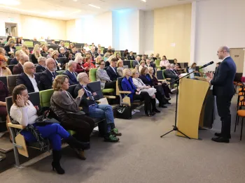 Sala konferencyjna, mężczyzna przy mównicy, publiczność siedzi na zielonych krzesłach.