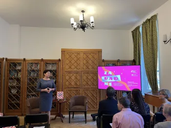 Kobieta przemawia do grupy ludzi w sali z drewnianymi regałami i ekranem z kolorową grafiką.