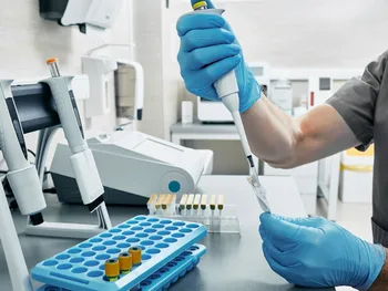 Osoba w rękawiczkach używa pipety w laboratorium, w tle sprzęt laboratoryjny.