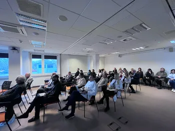 Grupa ludzi siedzących w sali konferencyjnej, niektórzy w białych fartuchach, patrzą na prezentację.