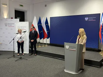 Trzy osoby przy mównicach w sali konferencyjnej, w tle flagi Polski i Unii Europejskiej.