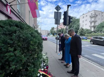 Cztery osoby stojące na chodniku, pochylone przed wieńcem z kwiatami.
