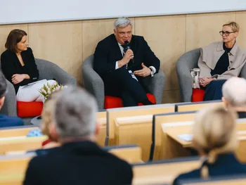 Trzy osoby siedzą na fotelach w sali konferencyjnej, jedna osoba mówi do mikrofonu.