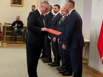 Mężczyzna wręcza dokument innemu mężczyźnie w eleganckiej sali z flagą w tle.