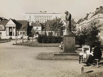 Pomnik Marii Skłodowskiej-Curie na postumencie, w tle budynki, na pierwszym planie osoba siedząca.
