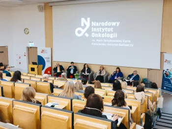 Sala konferencyjna, panel dyskusyjny, sześć osób siedzi na scenie, logo Narodowego Instytutu Onkologii na ekranie.