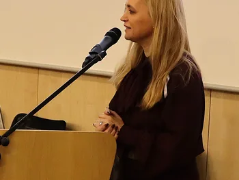 Kobieta przy mównicy mówi do mikrofonu, w tle ekran z logotypami i tekstem.