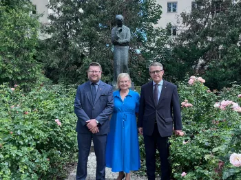 Trzy osoby stoją na ścieżce w ogrodzie, w tle pomnik i budynek.