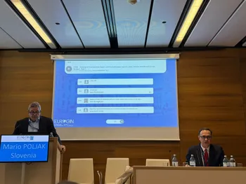 Dwóch mężczyzn na konferencji, jeden przy mównicy, drugi siedzi. Na ekranie prezentacja z tekstem.