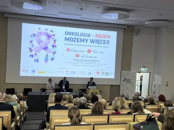 Sala konferencyjna, prelegenci na scenie, widownia siedzi. Na ekranie napis: 
