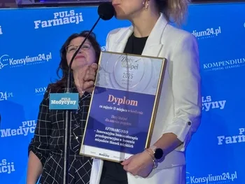 Kobieta w białej marynarce trzyma dyplom, obok kobieta w ciemnej sukience, w tle niebieskie logo Pulsu Medycyny.