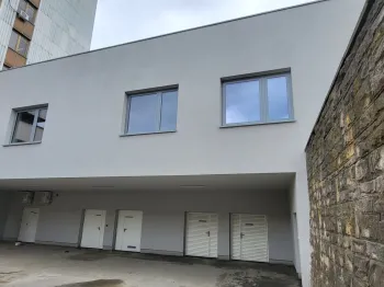 Budynek z białą fasadą, trzema oknami i garażami na parterze, obok kamienny mur.