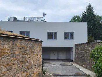 Budynek z białą fasadą, trzema oknami i dwoma drzwiami, otoczony murami i zielenią.