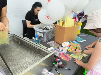 Dzieci przy stole z balonami, kredkami i materiałami plastycznymi na pikniku.