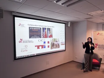 Kobieta prezentuje slajd z logotypami i zdjęciami, tekst: „Collaboration between the IC & the MSCI”.