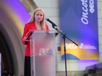 Kobieta w czerwonej marynarce przemawia przy mównicy na tle baneru 
