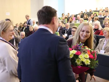 Osoba wręczająca bukiet kwiatów kobiecie w sali pełnej ludzi.