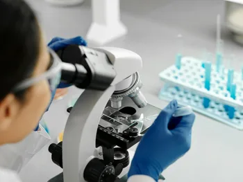 Osoba w rękawiczkach i okularach ochronnych używa mikroskopu w laboratorium.