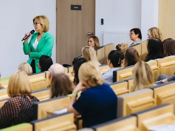 Kobieta w zielonej marynarce mówi do mikrofonu w sali wykładowej pełnej ludzi.