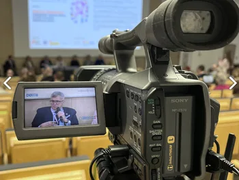Kamera na statywie w sali konferencyjnej, na ekranie projekcja prezentacji.