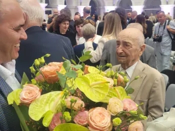 Osoby w eleganckich strojach, mężczyzna wręcza bukiet kwiatów starszemu mężczyźnie.