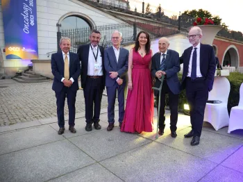 Grupa elegancko ubranych osób pozuje na zewnątrz przed budynkiem z łukami.