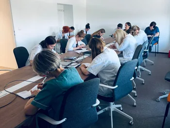 Grupa osób w białych fartuchach siedzi przy stołach w sali konferencyjnej, pisząc notatki.