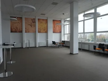Jasne pomieszczenie z dużymi oknami, stolikami i krzesłami, na ścianie obrazy w odcieniach brązu.