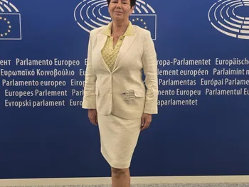 Kobieta w jasnym kostiumie stoi na tle niebieskiej ściany z logo Parlamentu Europejskiego.