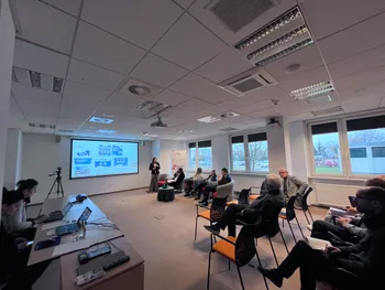 Sala konferencyjna z osobami siedzącymi, prezentacja na ekranie, osoba przemawia.