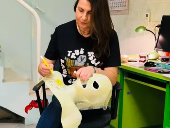 Kobieta siedzi na krześle i maluje żółtą maskę. W tle biurko i zielona szafka.