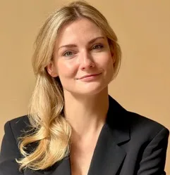 Kobieta z blond włosami w czarnej marynarce na beżowym tle.
