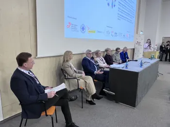 Siedem osób siedzi na krzesłach w sali konferencyjnej, obok stół z mikrofonami i butelkami wody.