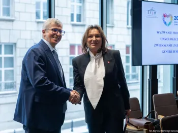 Mężczyzna i kobieta w formalnych strojach podają sobie ręce. W tle ekran z napisem o wyzwaniu onkologicznym.