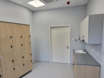 Pomieszczenie z szafkami na kluczyk, zlewem i szafką wiszącą, jasne ściany i sufit.