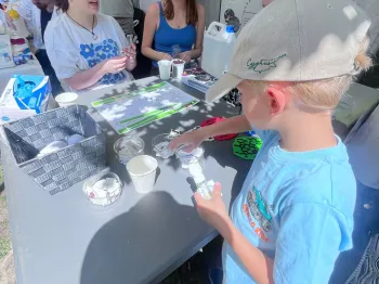 Dzieci i dorośli przy stoliku z materiałami edukacyjnymi na pikniku. Chłopiec w czapce trzyma kubek.
