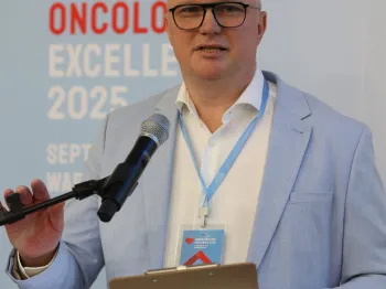 Mężczyzna w jasnym garniturze przemawia przy mikrofonie, trzymając notatnik. W tle napis: 