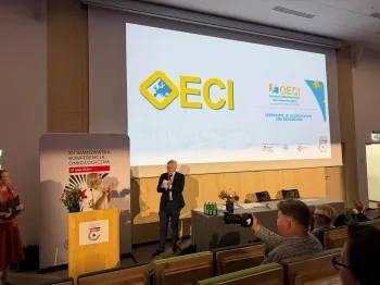 Osoby na sali konferencyjnej, mężczyzna wręcza certyfikat. Na ekranie logo ECI i OECI.