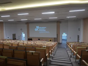 Duża sala wykładowa z drewnianymi krzesłami, ekranem projekcyjnym i napisem: Narodowy Instytut Onkologii.
