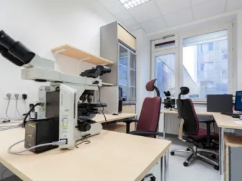 Laboratorium z mikroskopami, biurkami i krzesłami obrotowymi, jasne ściany, duże okno.
