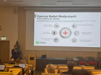 Mężczyzna przemawia w sali wykładowej, obok choinka. Na ekranie prezentacja Agencji Badań Medycznych.