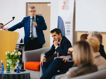 Mężczyzna w garniturze przemawia przy mównicy, obok siedzący uczestnicy konferencji, w tle baner.