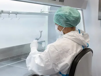 Osoba w białym kombinezonie ochronnym i zielonej czepce pracuje w laboratorium przy dygestorium.