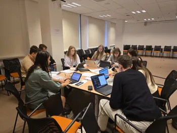 Grupa osób siedzi przy stole w sali konferencyjnej, pracując na laptopach.