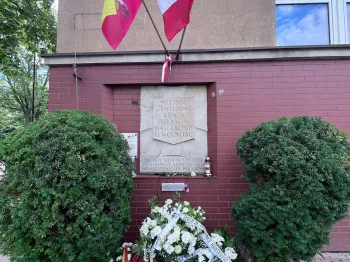 Tablica pamiątkowa z wieńcami i zniczami, nad nią dwie flagi na ścianie budynku.
