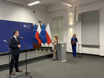 Trzy osoby na podium, flagi Polski i UE w tle, mikrofony, mężczyzna w garniturze, kobiety w formalnych strojach.