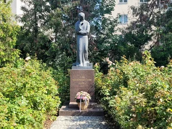 Pomnik postaci w ogrodzie, otoczony krzewami i kwiatami, z koszem kwiatów u podstawy.
