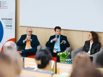 Trzy osoby siedzą na scenie podczas panelu dyskusyjnego, obok roll-up z logo i napisem „Rzetelność w badaniach klinicznych”.